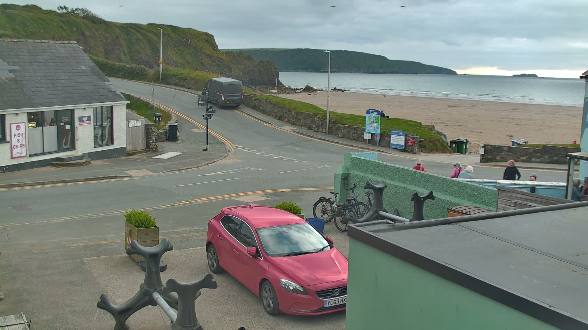 A pink car is parked near a road that leads to a beach with calm waves under an overcast sky.