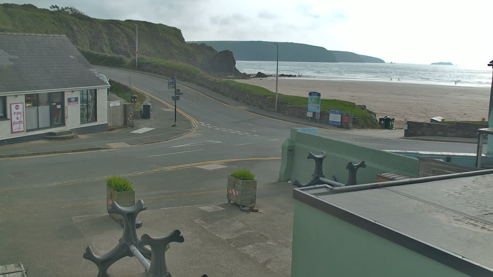 A coastal road curves toward a sandy beach under an overcast sky, with a small building and decorative anchors visible in the foreground.