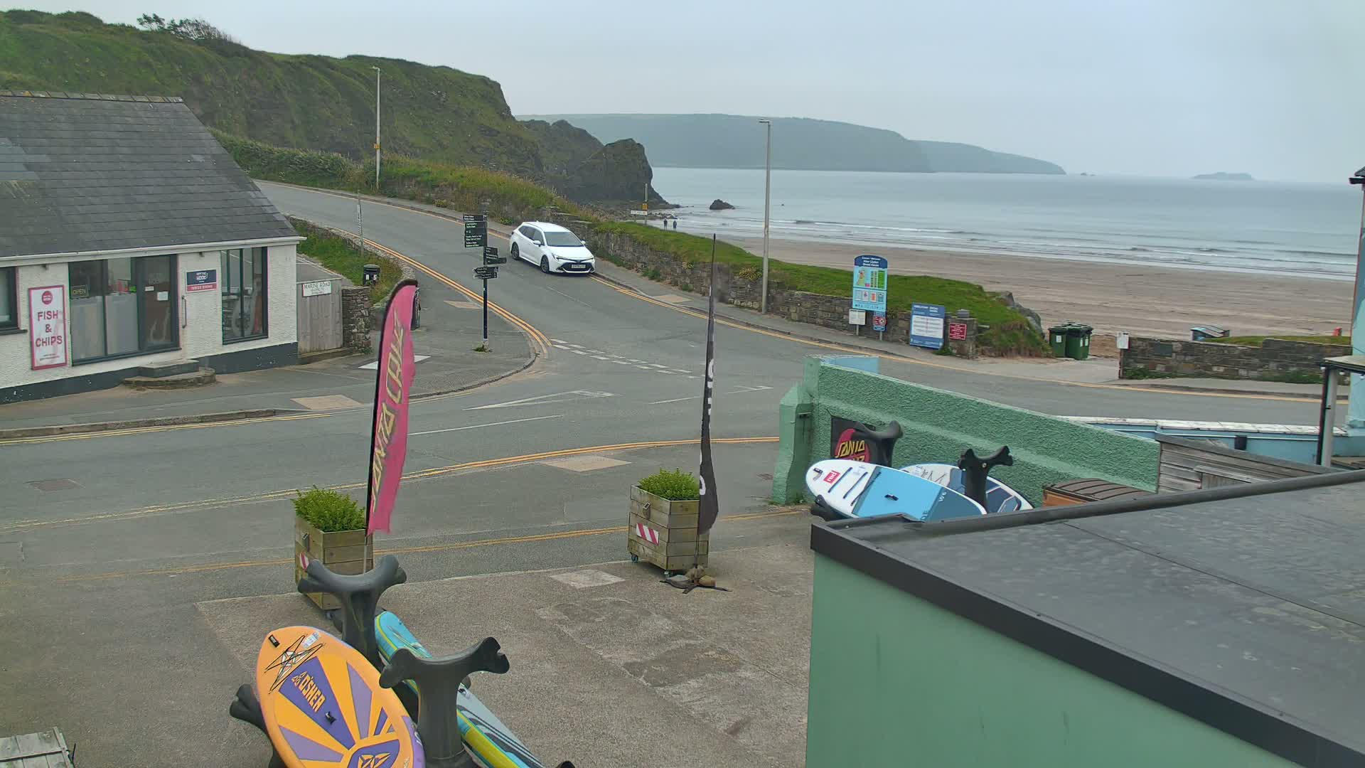 A seaside town street scene on an overcast day shows a white car turning onto a road that leads to a sandy beach with calm waves.
