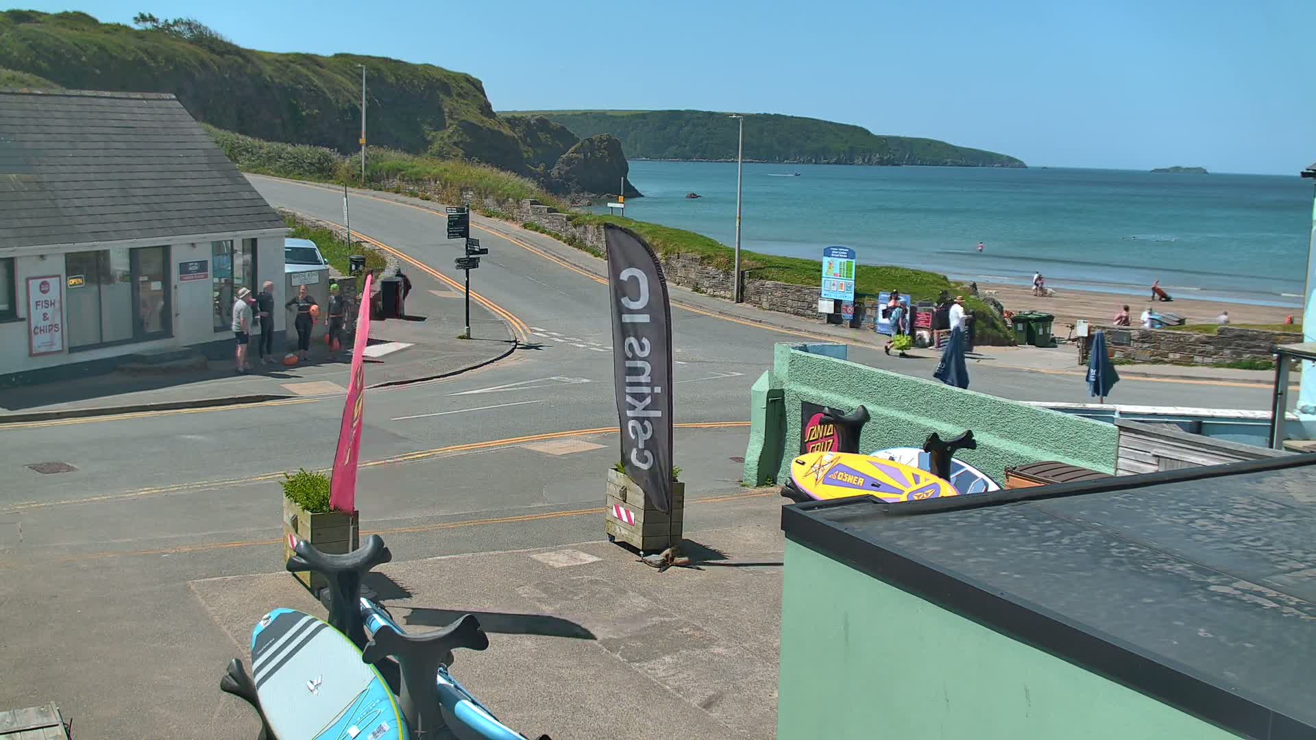 A sunny coastal town scene shows a road intersection near a beach with people walking and several paddleboards, one of which is bright blue.