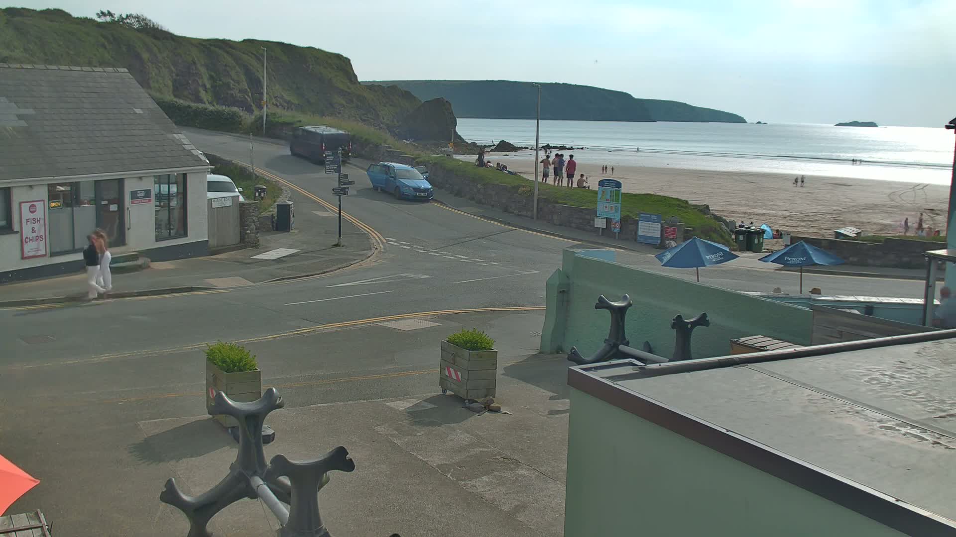 A seaside town scene on a sunny day shows a road intersection near a sandy beach with people and cars, and a building selling fish and chips.
