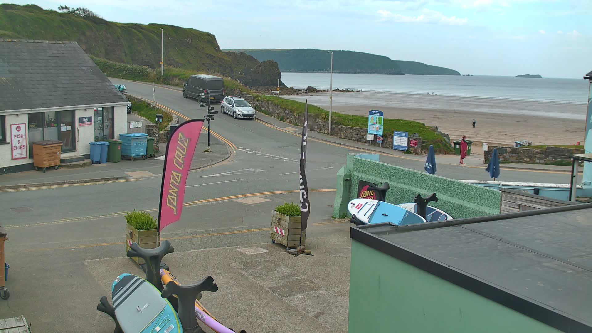 A seaside town scene on a partly cloudy day shows a road curving toward a sandy beach with people strolling, vehicles parked and driving, and surfboards leaning against a building.