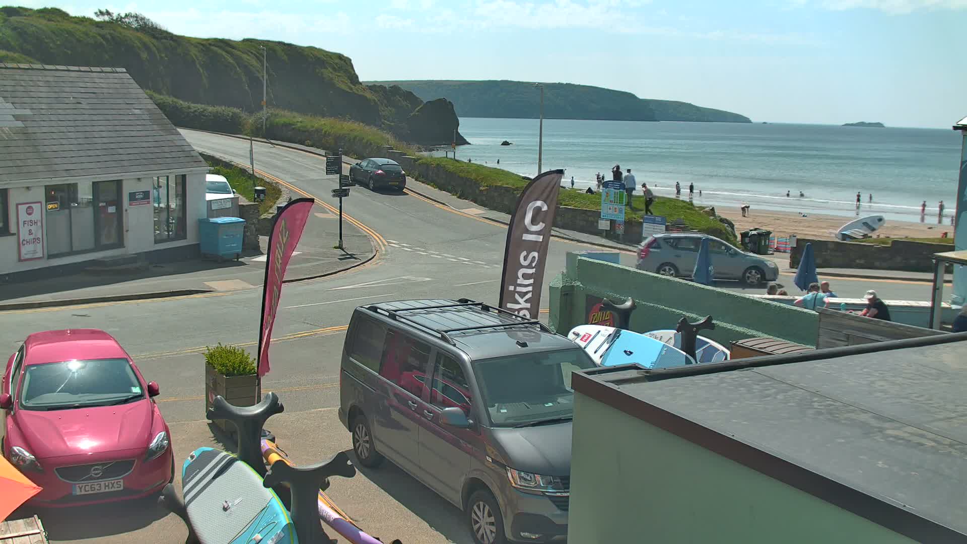 A sunny coastal scene shows a road curving toward a beach with people on the sand, several parked cars near buildings, and surfboards leaning against a structure.