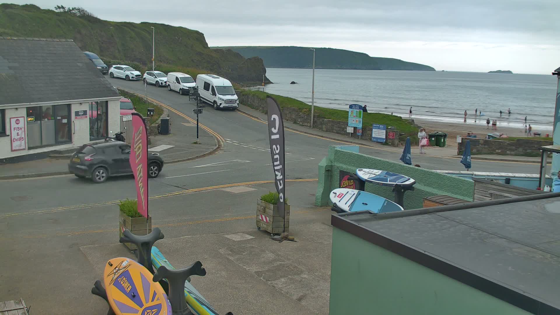 A coastal road scene with overcast skies shows parked cars near a shop, a beach with people, and several paddleboards leaning against a building.
