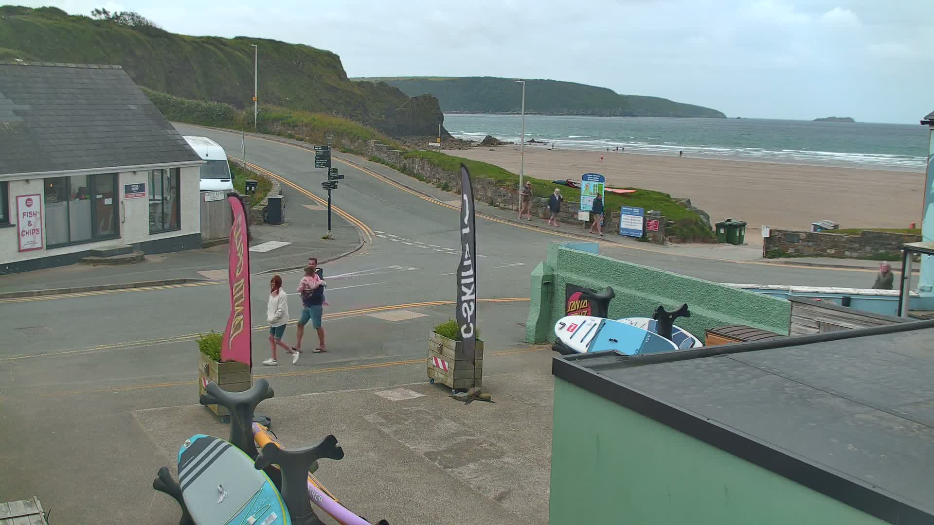 A street scene overlooking a sandy beach with gentle waves under an overcast sky, showing a few people walking near a road intersection and some surfboards leaning against a building.