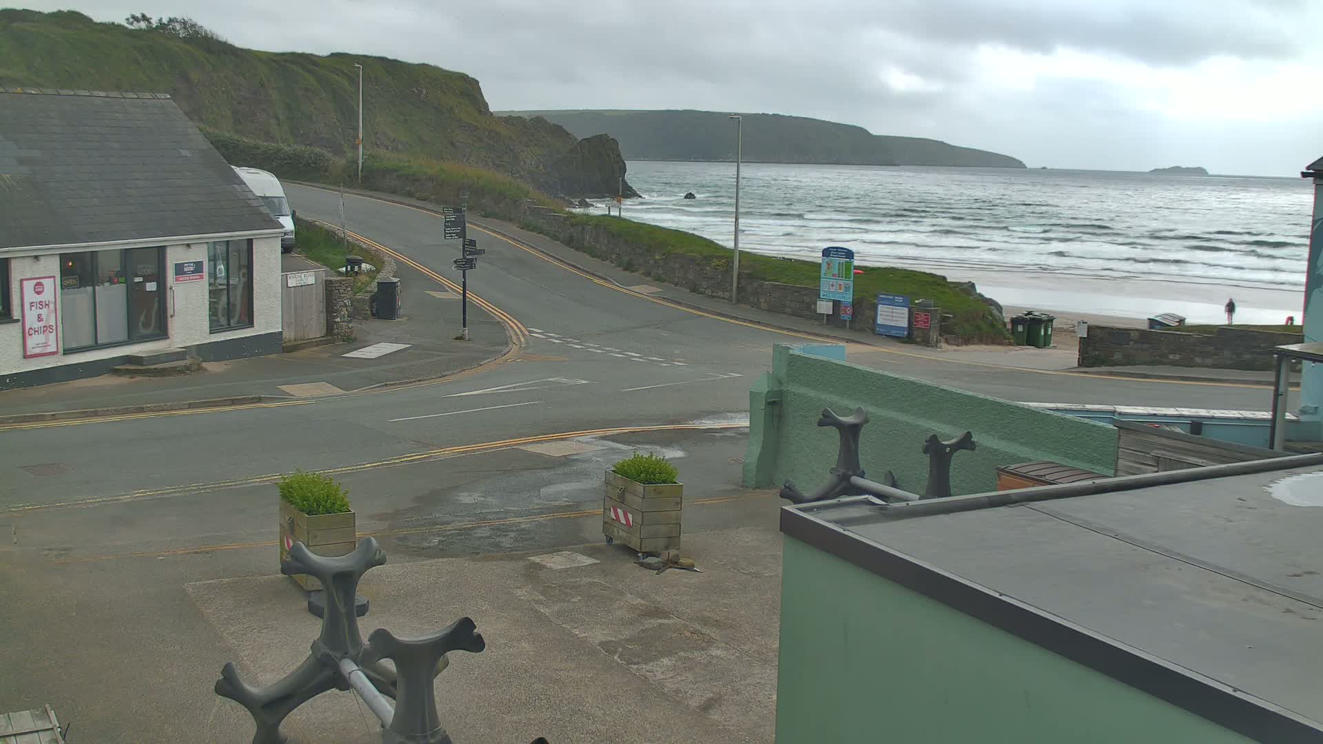 A seaside road intersects near a fish and chips shop, overlooking a beach with choppy waves under a cloudy sky.
