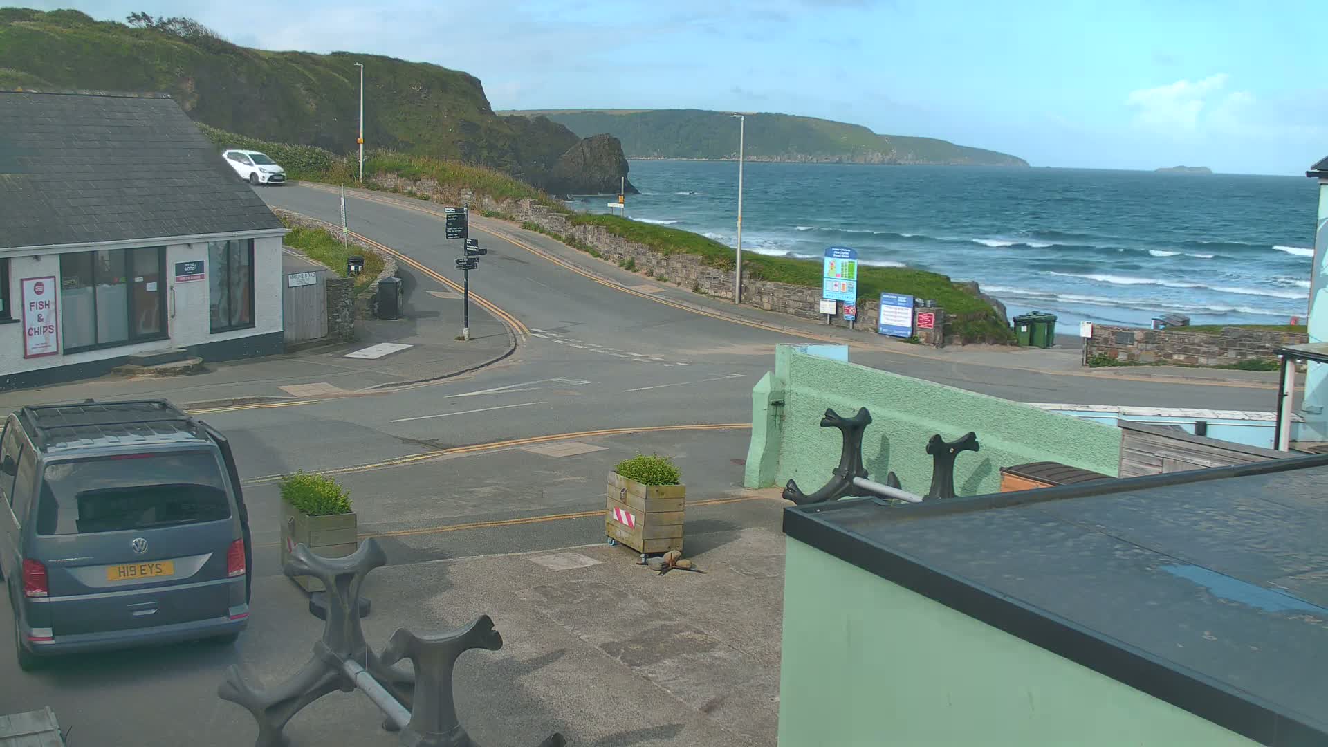 A coastal road scene shows a partially cloudy sky, a wavy ocean with small waves, and several vehicles near a building.