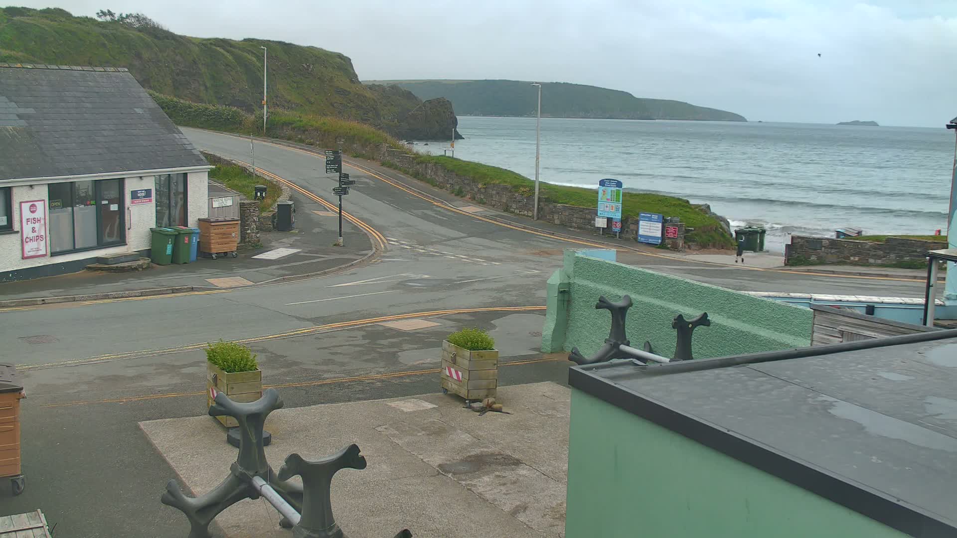 A coastal road curves toward a beach with small waves under an overcast sky, with a building and several waste bins visible.