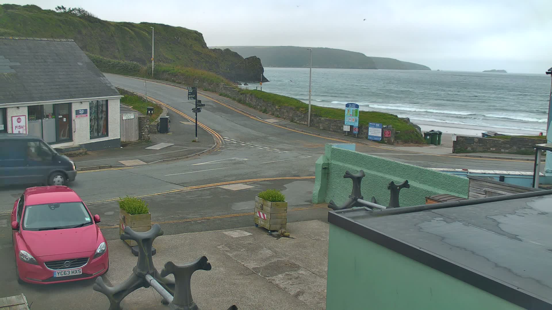 A pink car is parked near a road intersection overlooking a beach with waves under an overcast sky.