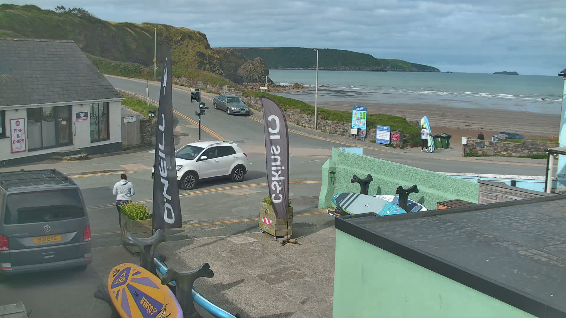 A coastal scene on a partly cloudy day shows a beach with calm waves, cars on a road near buildings, and a person carrying a surfboard.