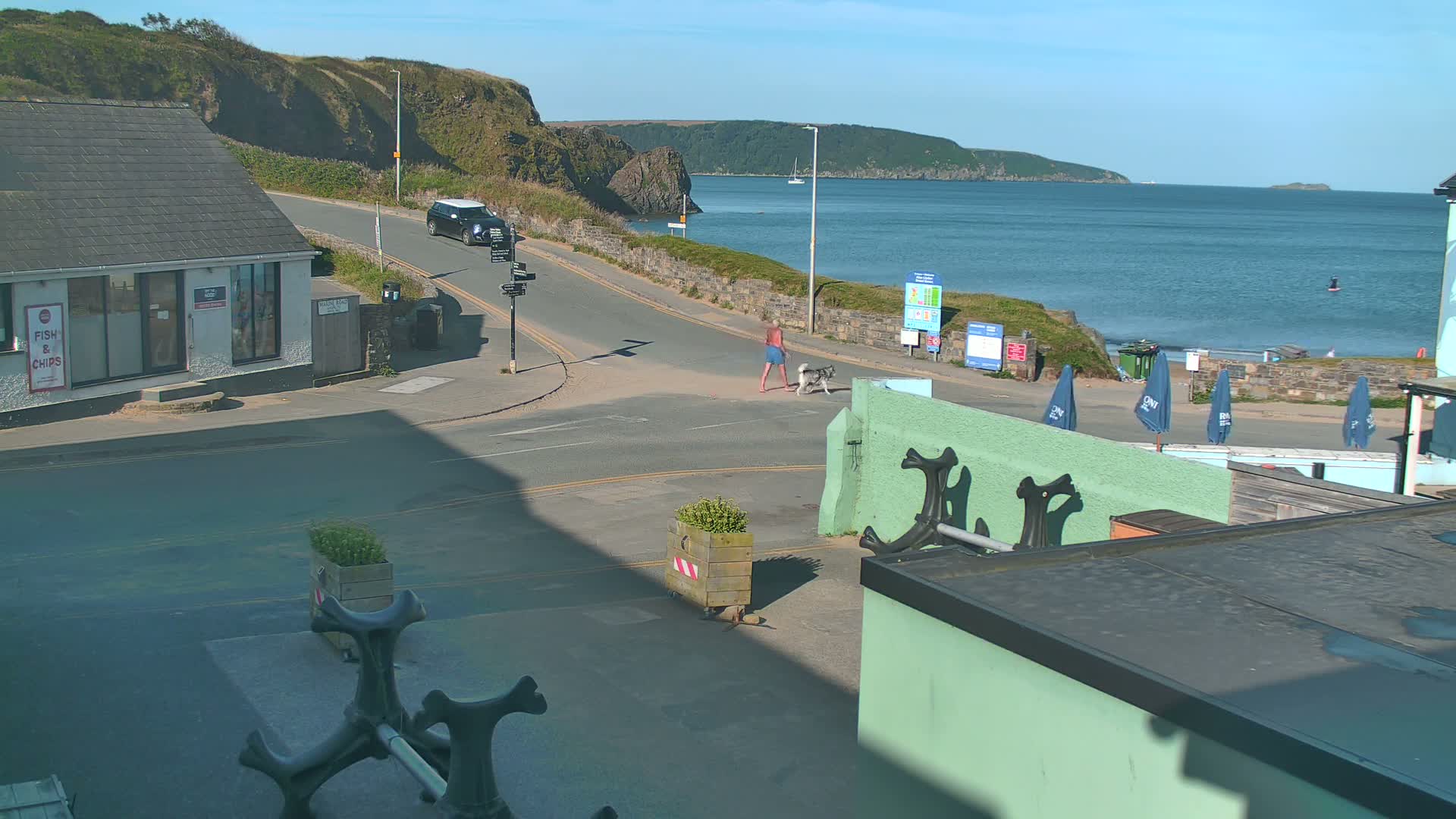 A man walks his dog across a road near a calm ocean on a sunny day, with a small building and several closed umbrellas visible nearby.