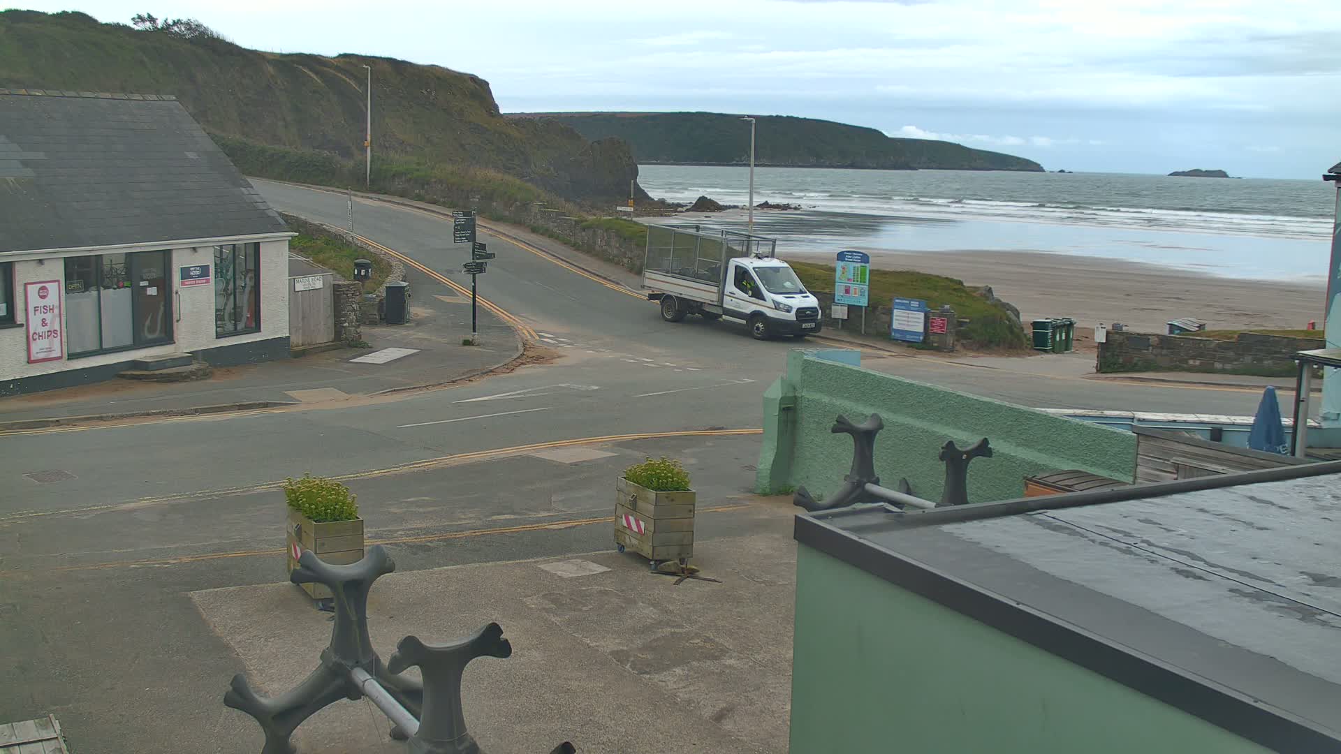 A white van is turning onto a road near a beach with small waves under an overcast sky.