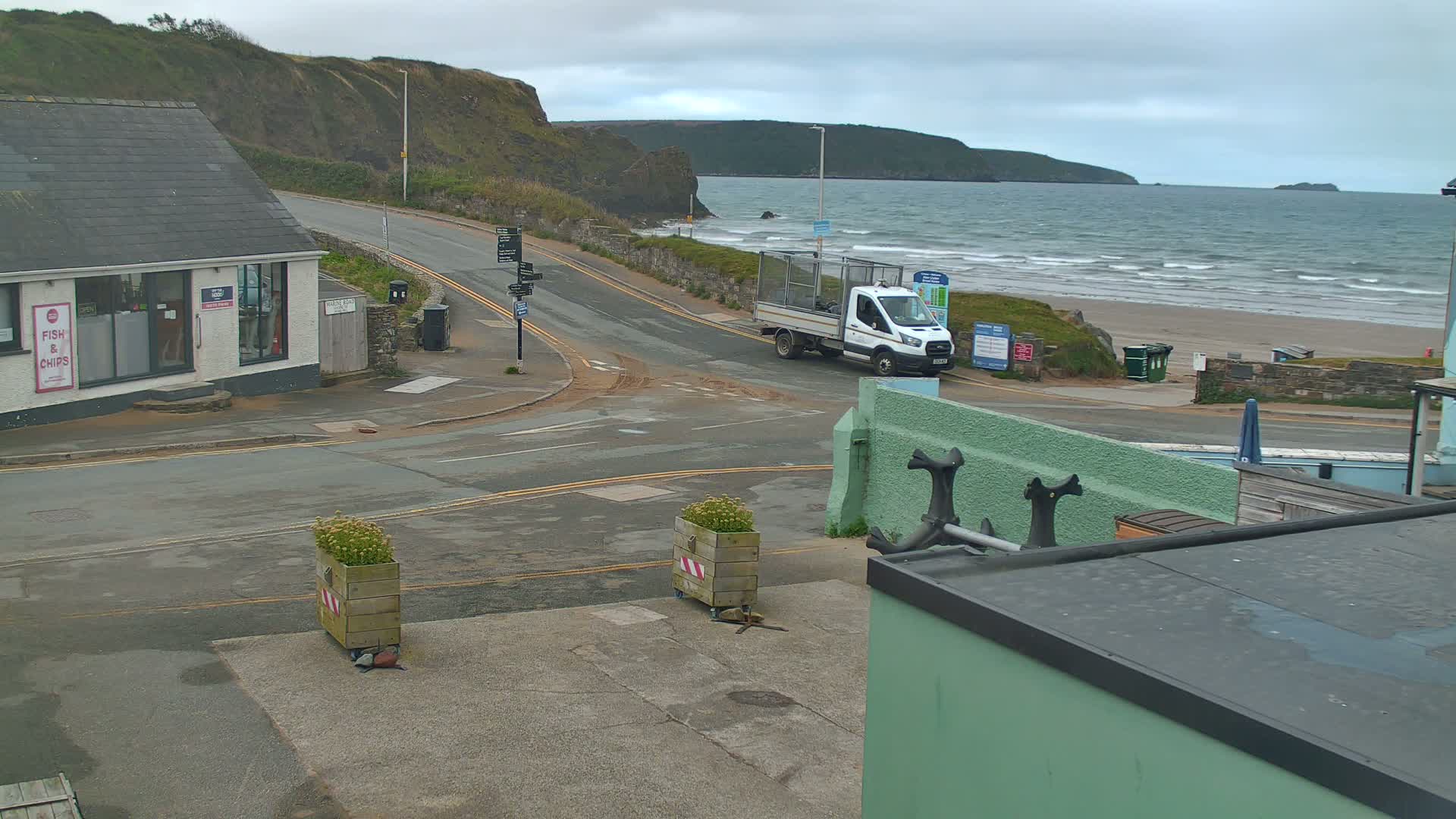 A white delivery truck is parked near a road intersection overlooking a beach with wavy water under an overcast sky.