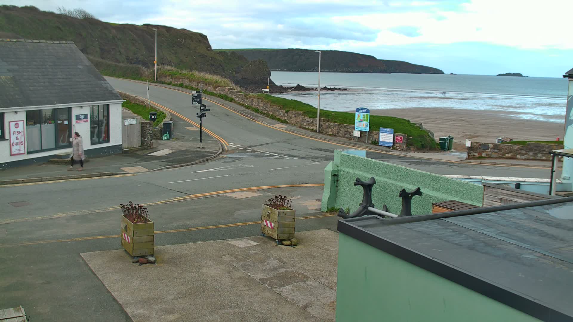 On an overcast day, a person walks past a roadside building on a coastal road overlooking a wide, sandy beach and the sea, backed by green cliffs.