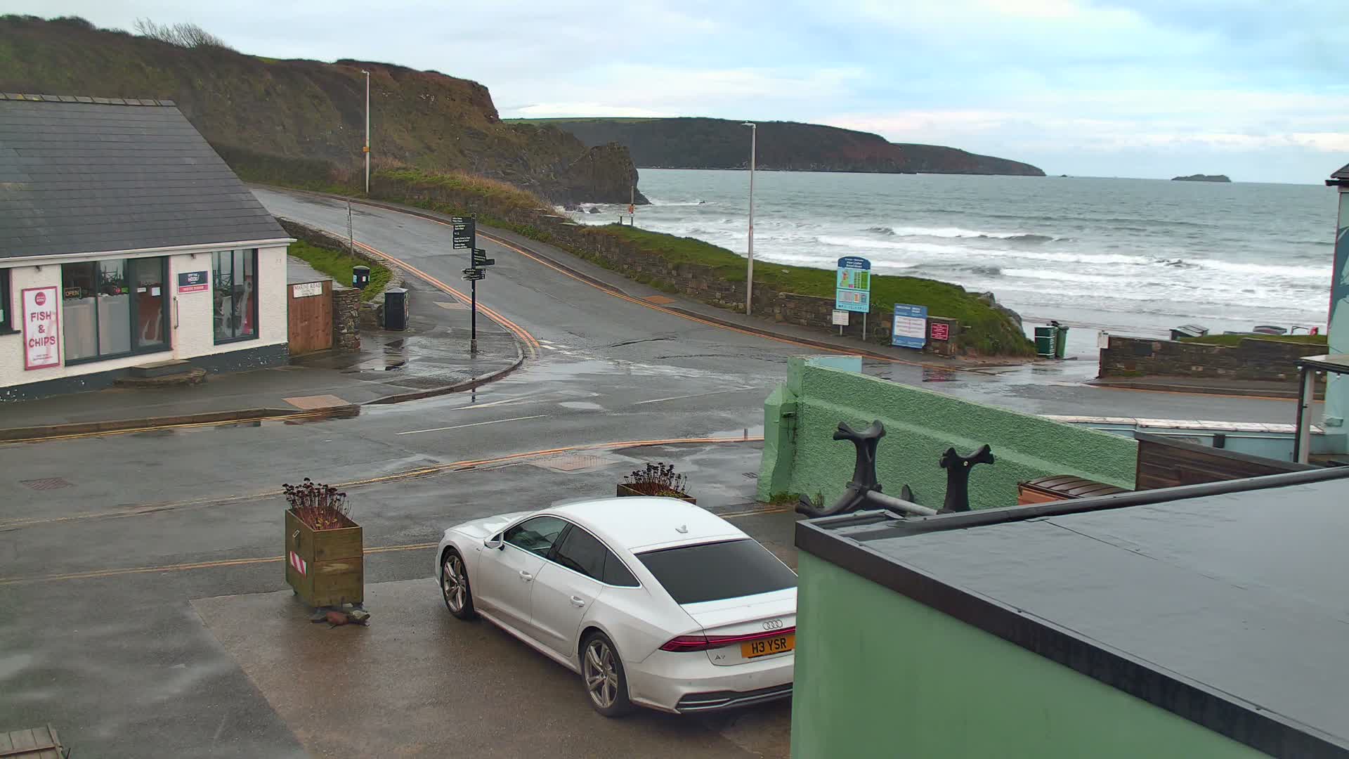 A white car is parked on a wet street in a coastal village, with a small shop on the left and a choppy gray sea breaking against the shore with distant cliffs under an overcast sky, indicating a damp and cloudy day.