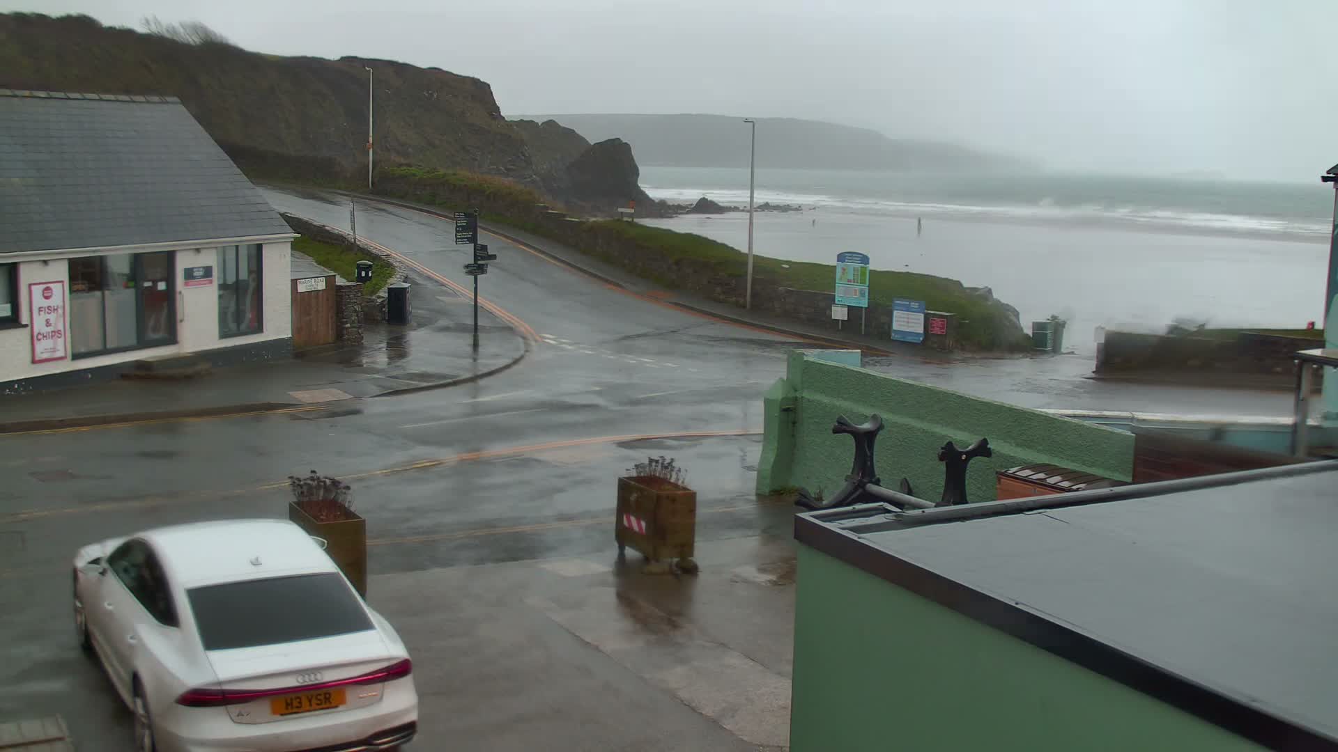 A coastal scene on a gloomy, wet, and overcast day shows a white car on a damp road beside a building, with rough waves breaking on a beach against a backdrop of green cliffs.