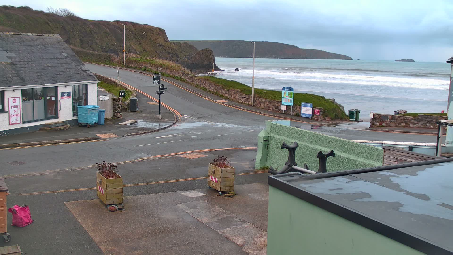 A grey, overcast day reveals a wet coastal road curving past a fish and chips shop towards a beach with crashing waves, backed by steep cliffs and distant headlands.