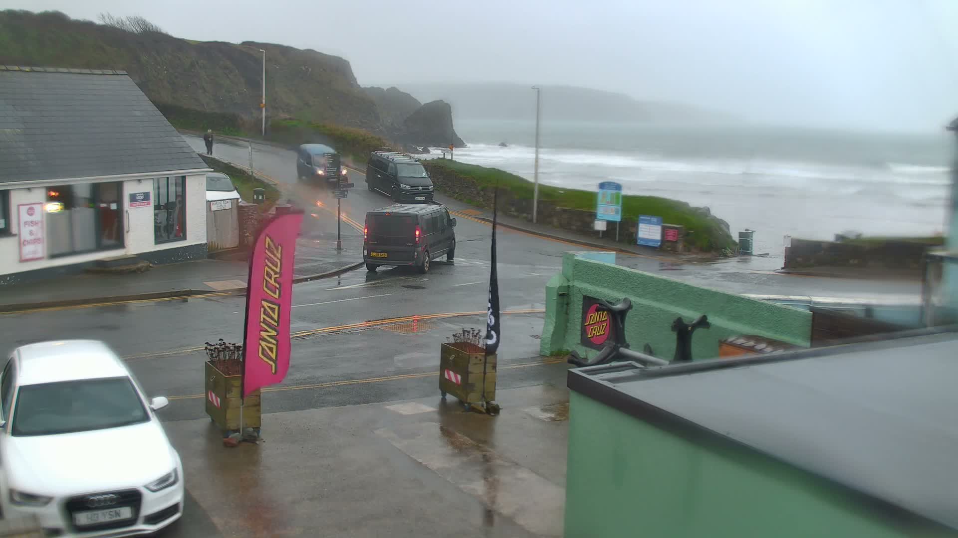 A grey, overcast day reveals a wet coastal road curving past a fish and chips shop towards a beach with crashing waves, backed by steep cliffs and distant headlands.