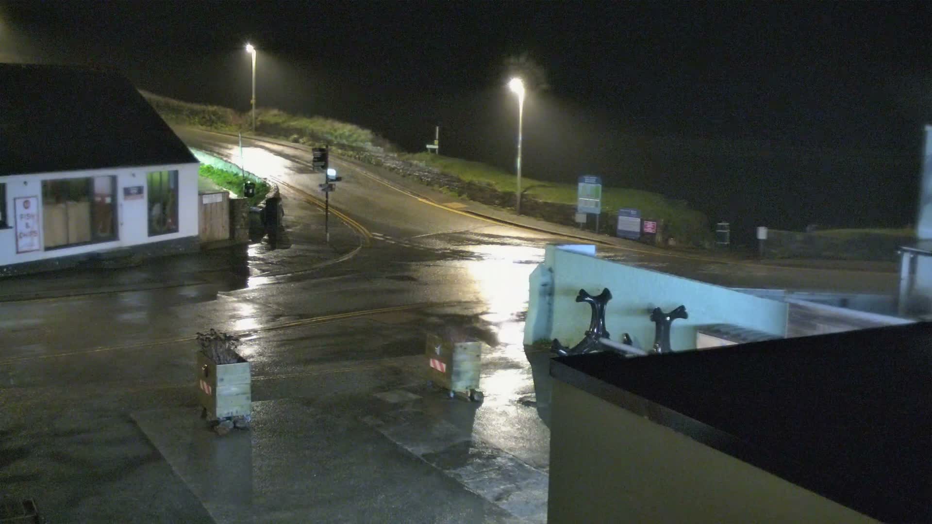 A grey, overcast day reveals a wet coastal road curving past a fish and chips shop towards a beach with crashing waves, backed by steep cliffs and distant headlands.