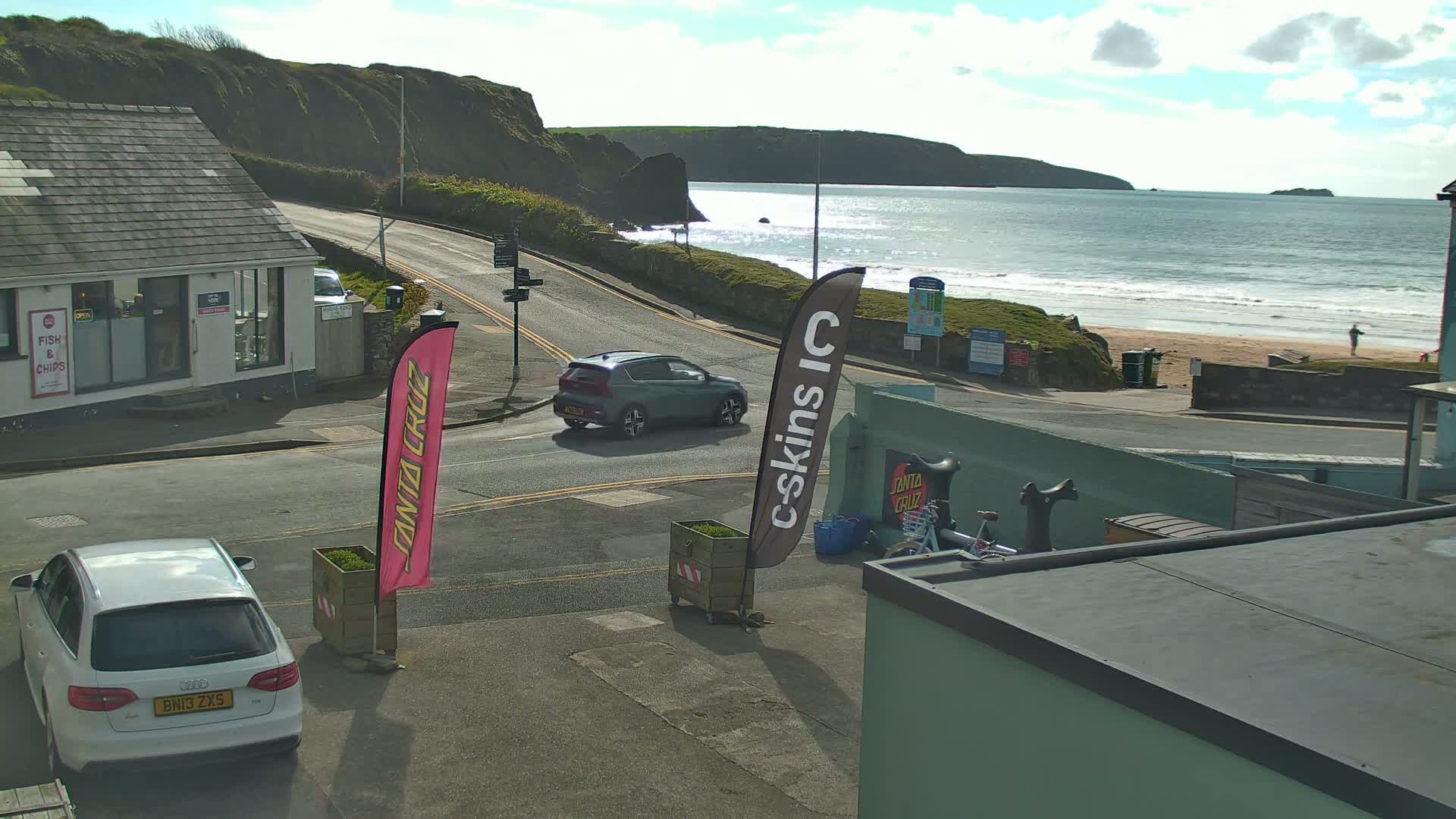 A grey, overcast day reveals a wet coastal road curving past a fish and chips shop towards a beach with crashing waves, backed by steep cliffs and distant headlands.