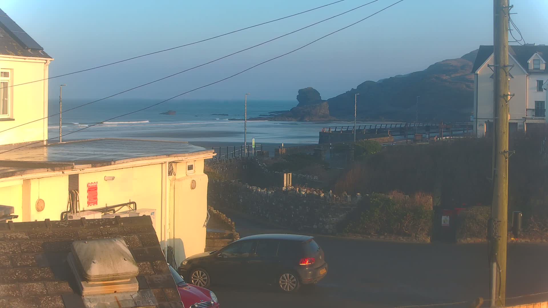 A view of a beach with small waves under a clear, bright sky, seen from between buildings, with a dark-colored car parked on a street in the foreground.