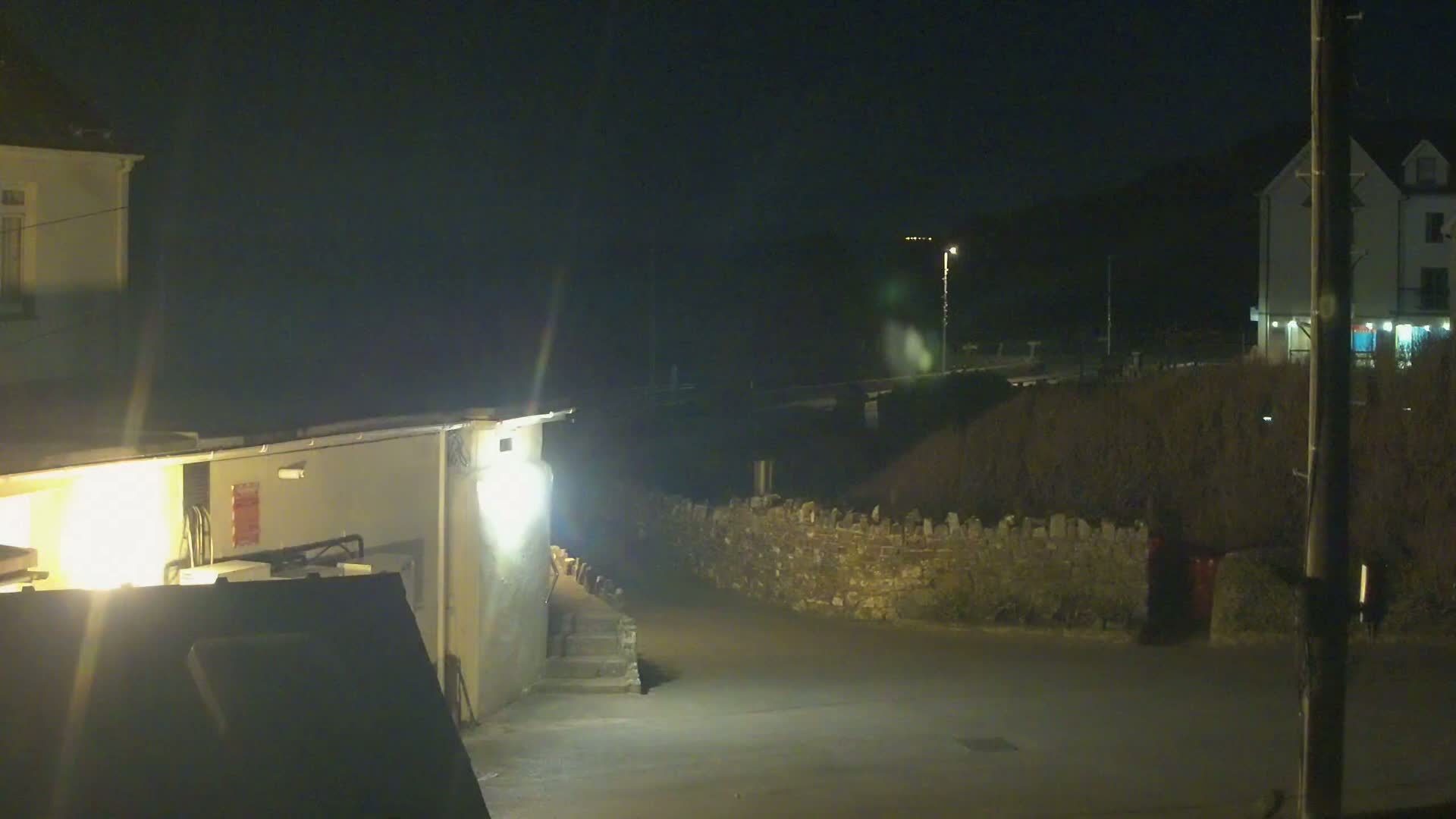 A nighttime scene shows a paved area between two buildings, one of which has exterior lighting illuminating a short set of steps, all under a dark sky.