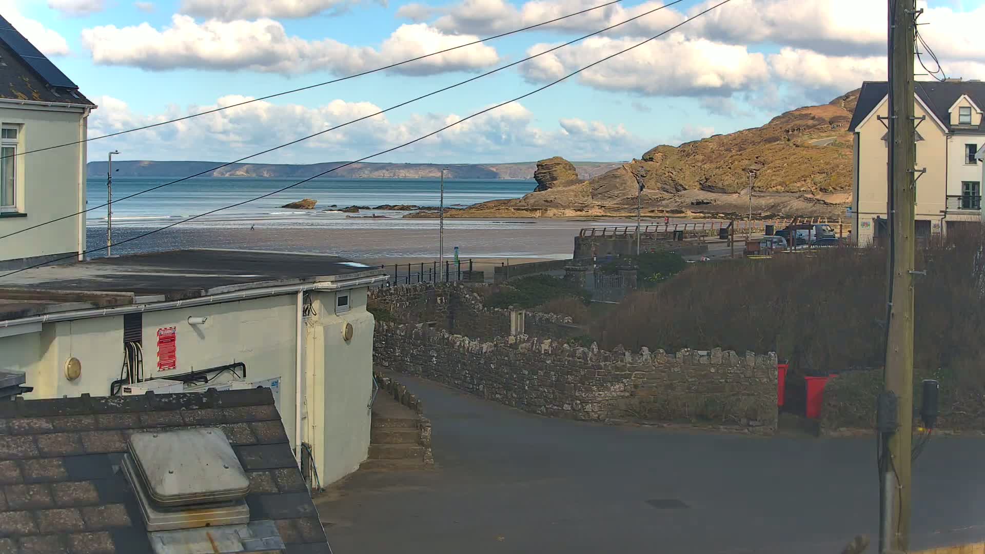A partly cloudy day reveals a coastal scene with a sandy beach, calm ocean waves, and rock formations, viewed from a street between buildings.