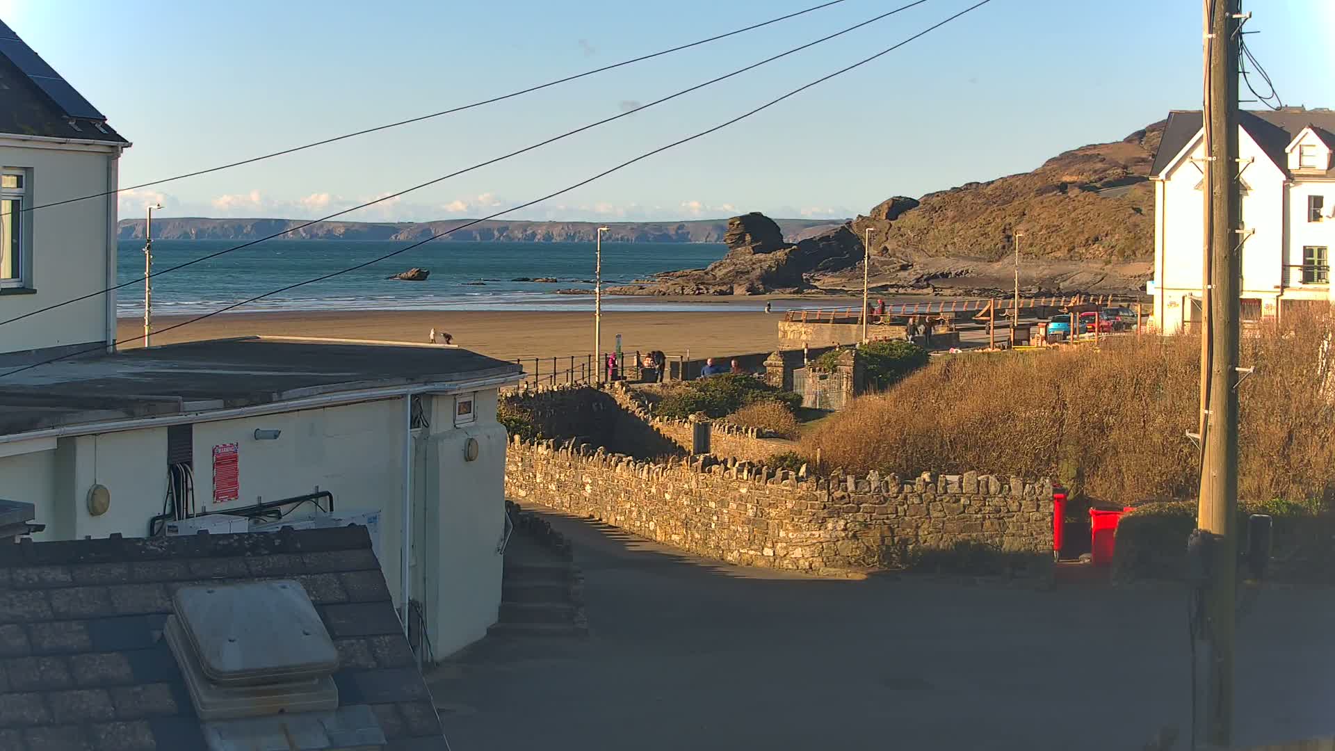 A sandy beach with a few people on it is visible from a vantage point between buildings, under a mostly clear, sunny sky.