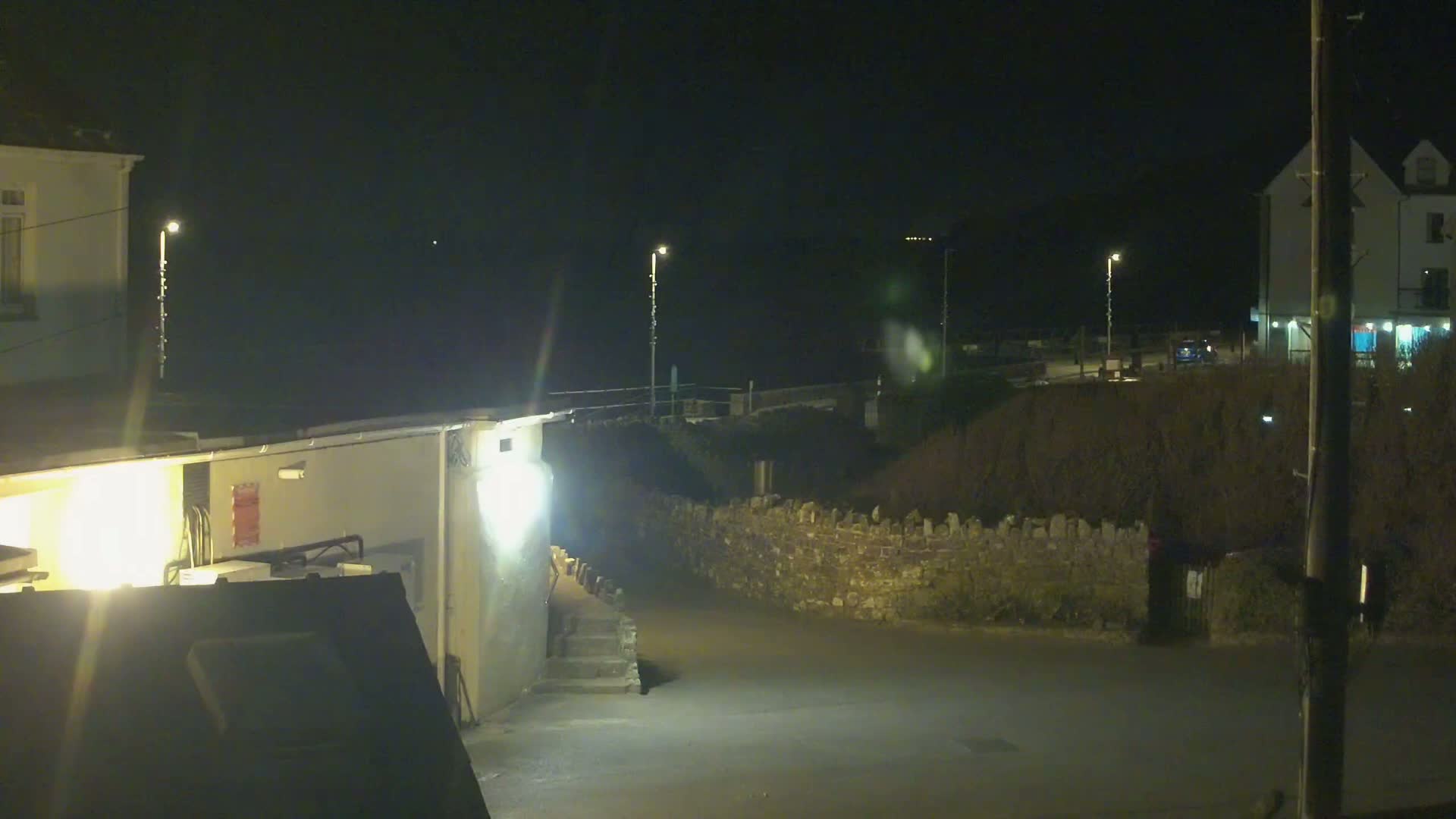 A nighttime view of a dimly lit street scene with buildings, a low stone wall, and sparsely vegetated areas, under dark skies.