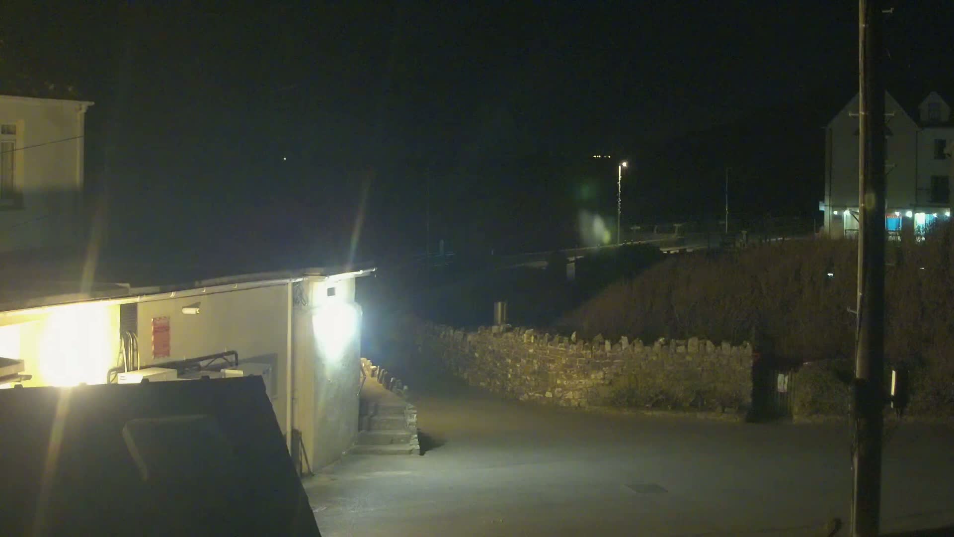 At night, a dimly lit street scene shows buildings, a low stone wall, and a sparsely vegetated area under dark skies.