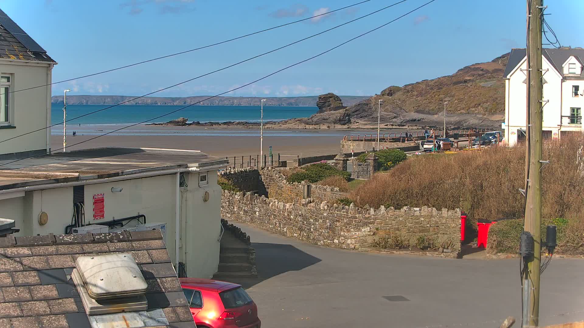 A sunny seaside scene shows a sandy beach with calm water, a stone wall, buildings, and a red car partially visible.