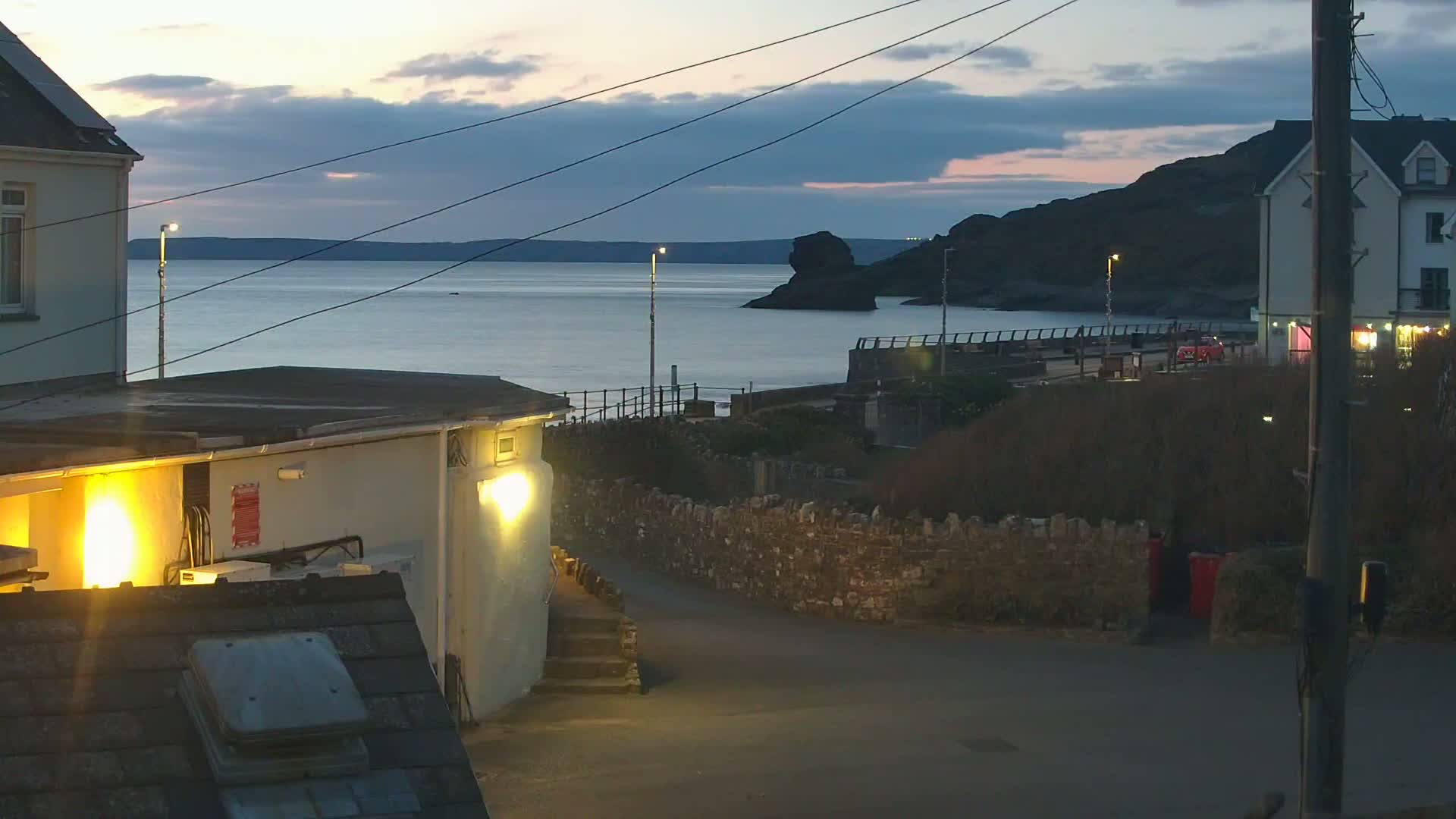 A calm, twilight sea is visible beyond a coastal road and buildings, with a stone wall separating the road from the water's edge.
