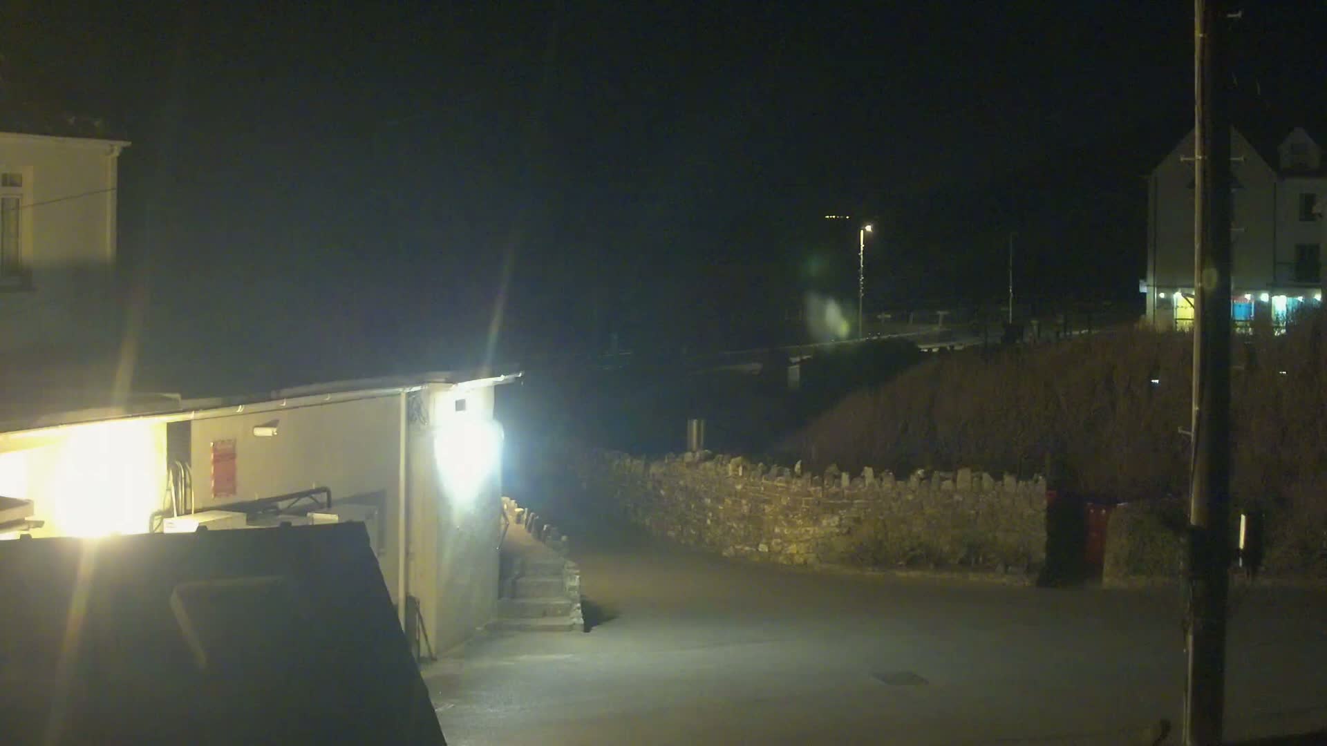 A nighttime view of a dimly lit street scene with buildings, a low stone wall, and a streetlight.