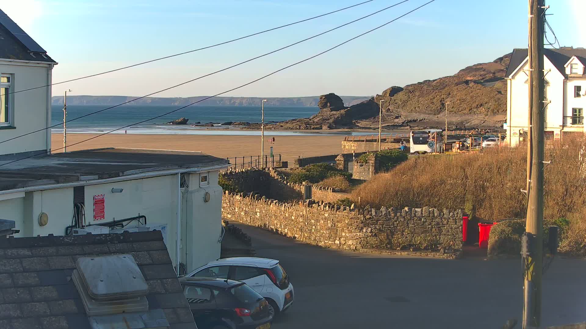 A sunny view overlooking a sandy beach, rocky coastline, and parked cars, with buildings and a stone wall in the foreground.