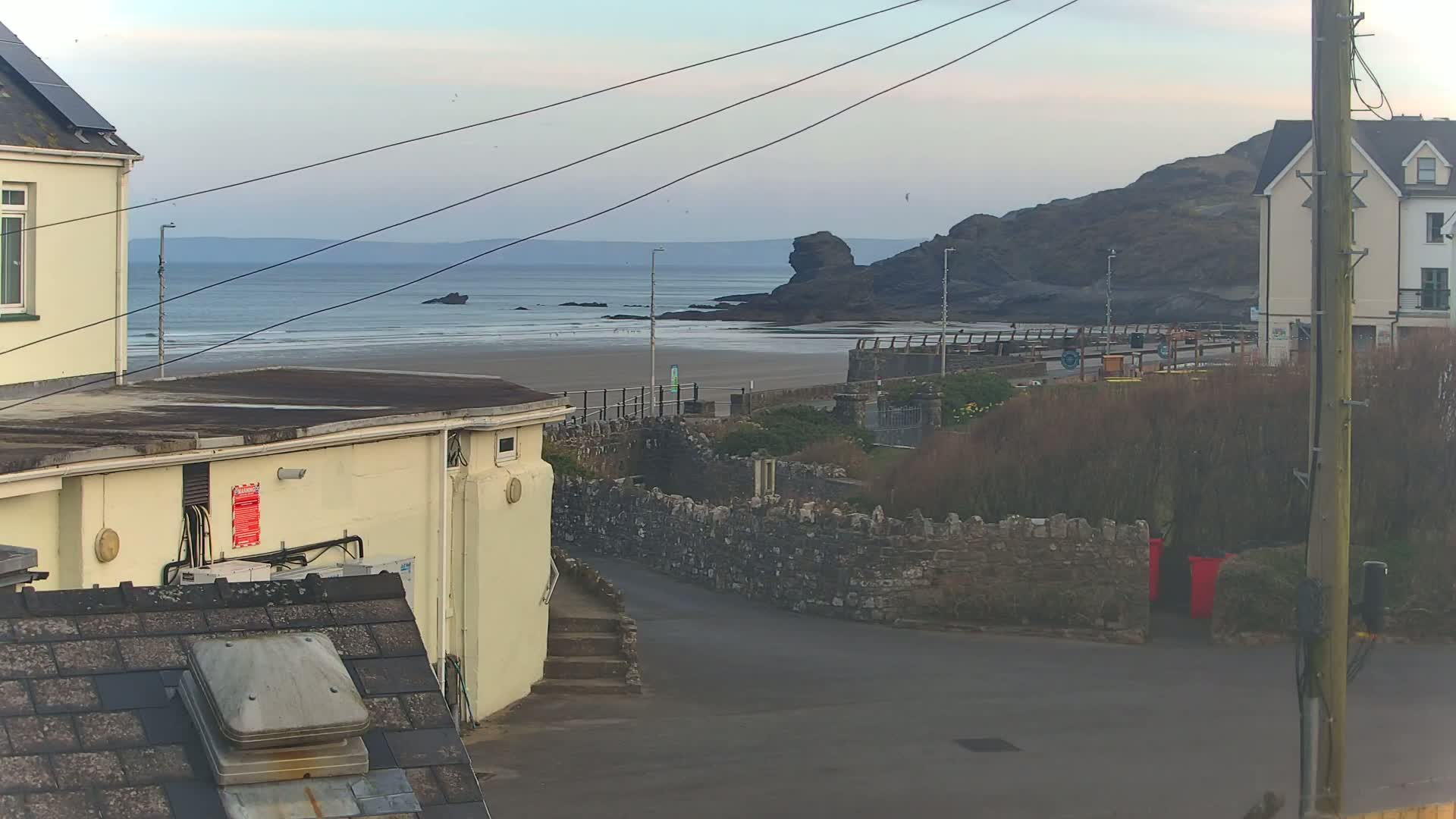 A view from a rooftop shows a sandy beach, calm ocean, rocky shoreline, buildings, and a stone wall under an overcast sky.