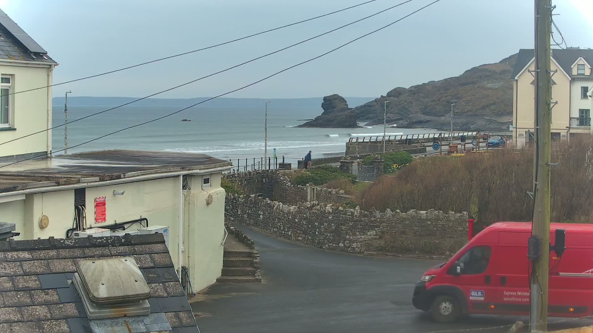 A view of a coastal town on an overcast day, showing a beach with small waves, a rocky outcrop, buildings, and a red delivery van on a road.