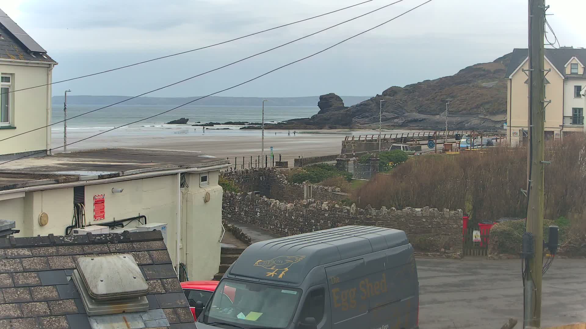 A gray van is parked near buildings overlooking a sandy beach with a rocky outcrop and overcast skies.