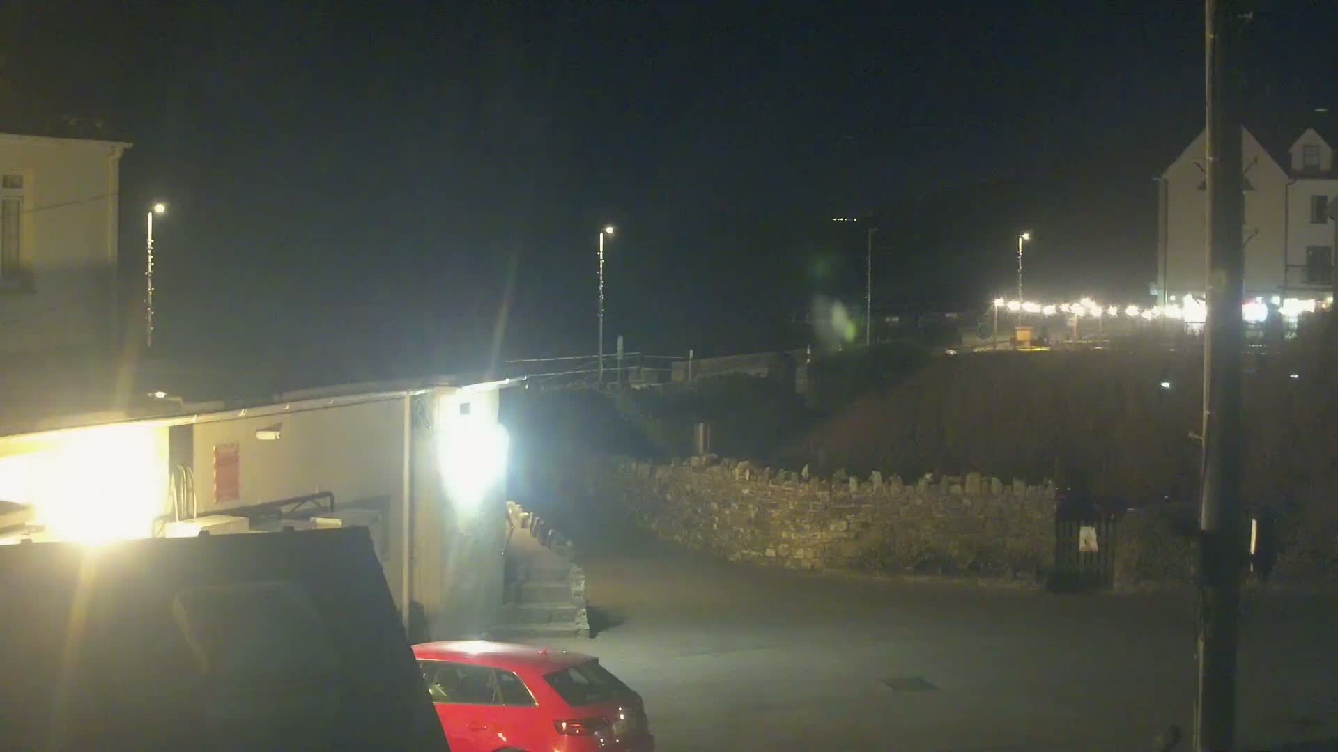 A nighttime view of a parking lot near buildings and a low stone wall, illuminated by streetlights.