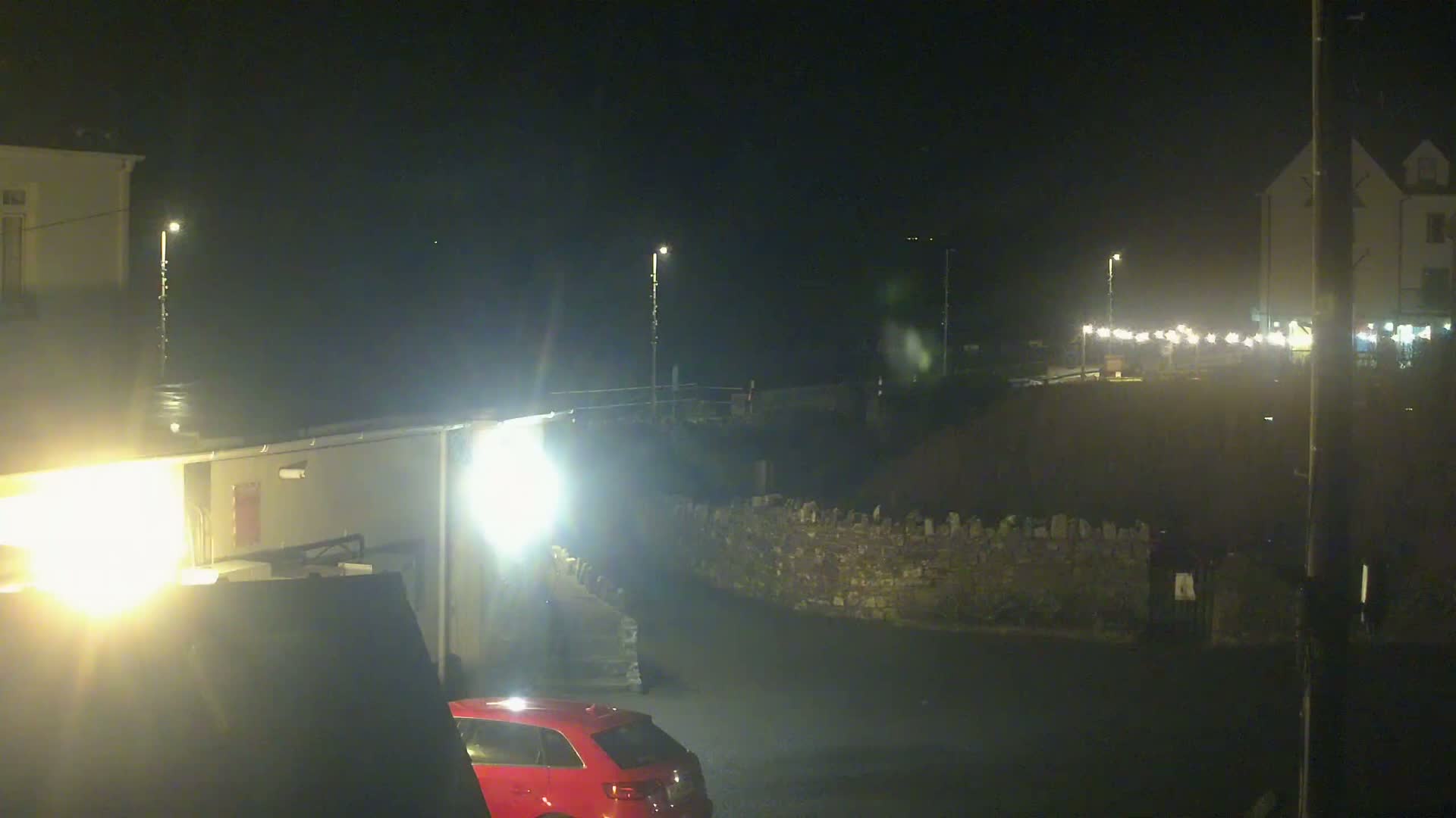 A nighttime scene shows a red car parked near buildings and a low stone wall, with bright lights illuminating the area.