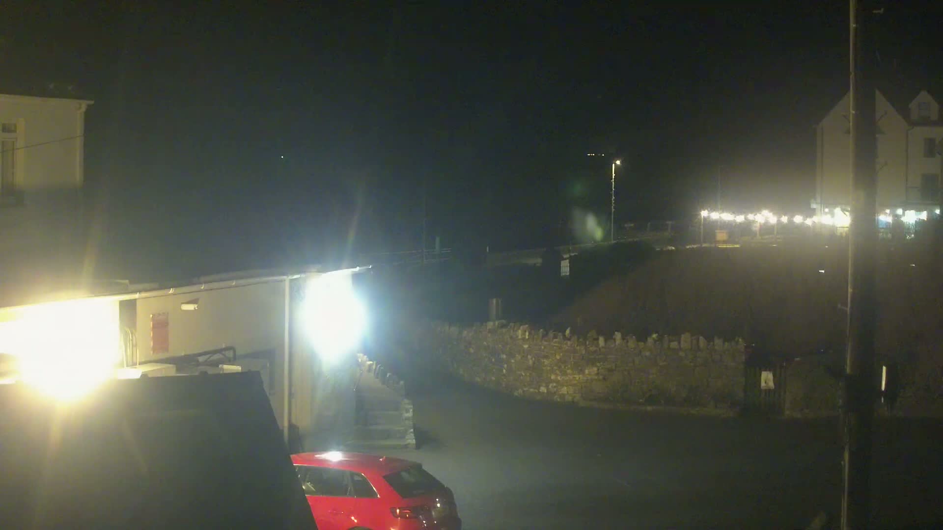 A red car is parked near a stone wall at night, with brightly lit buildings and a string of lights in the background under a dark sky.