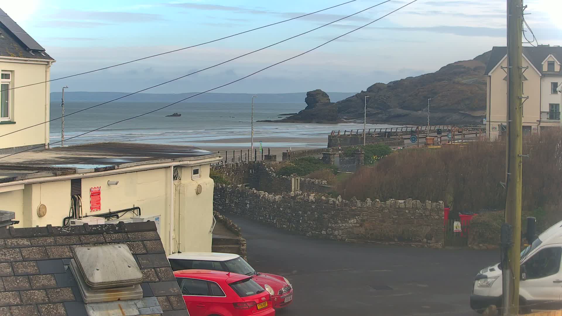 A view of a sandy beach with calm ocean waves under a cloudy sky, seen from a coastal town with parked cars and buildings.