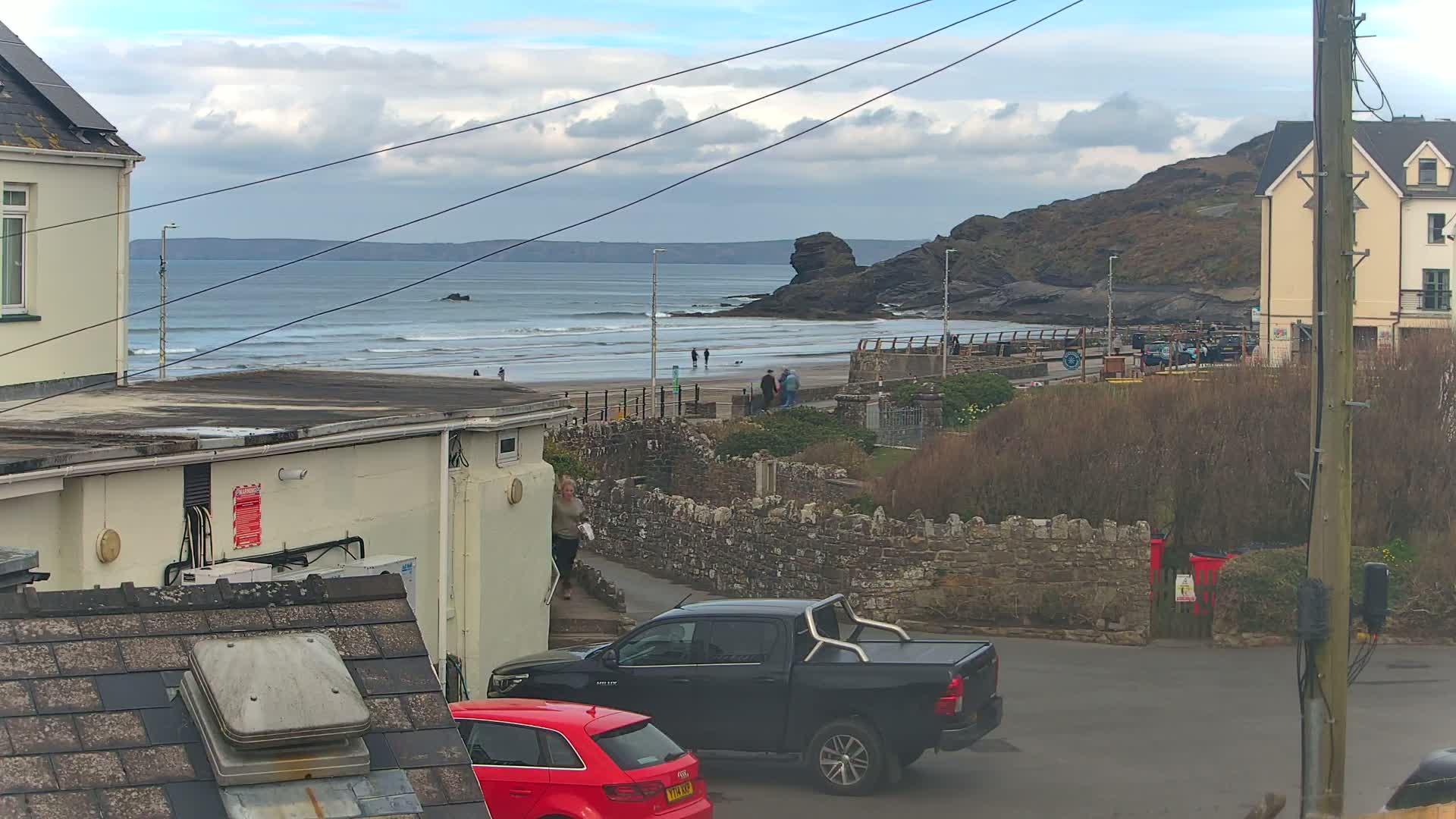 A view of a beach with small waves under a partly cloudy sky, seen from a residential area with parked cars and buildings.
