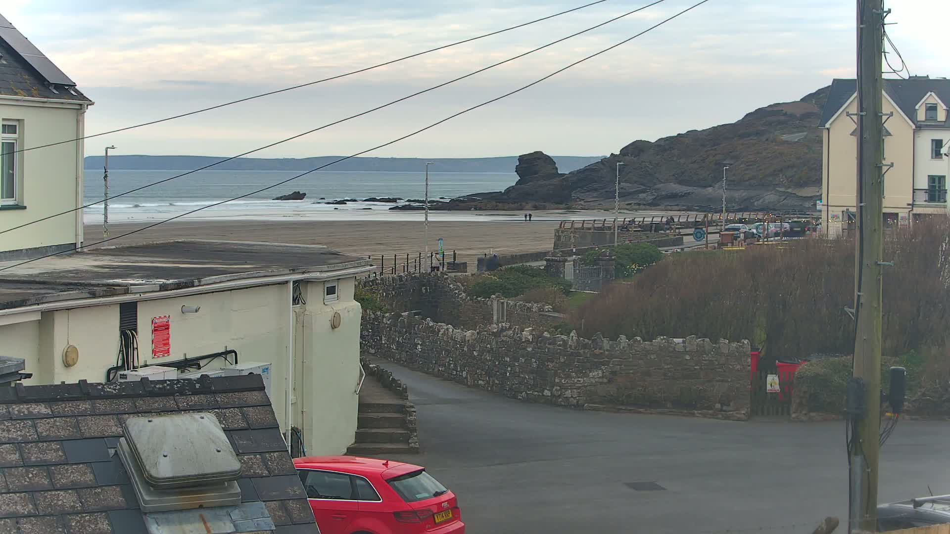 A view of a sandy beach with calm waves under an overcast sky, seen from between buildings on a small road leading down towards the shore.