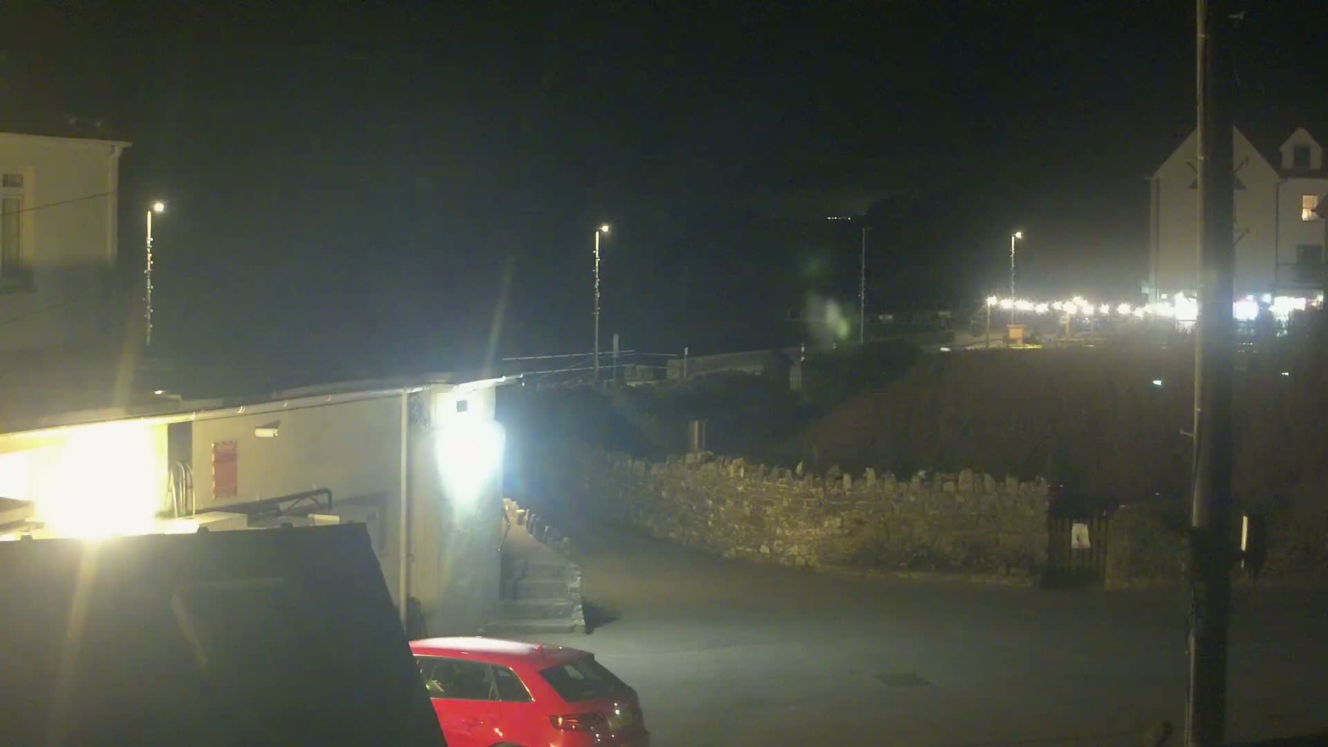 A nighttime scene shows a red car parked in a small lot next to a stone wall, with buildings and lights visible in the background under a dark sky.