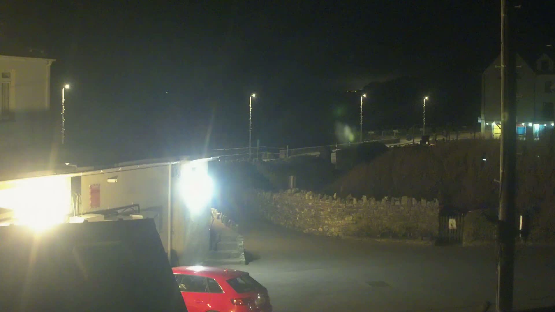 A nighttime scene shows a red car parked near a building, a stone wall, and several lit streetlights.