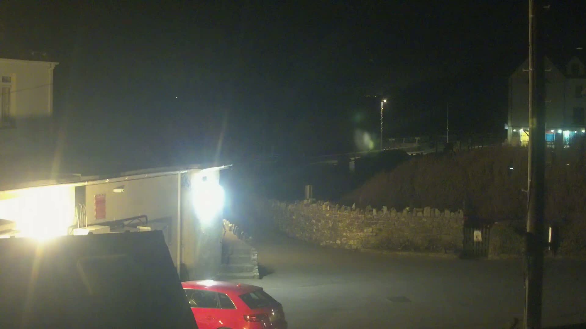 A red car is parked in a dimly lit area at night near buildings and a low stone wall.