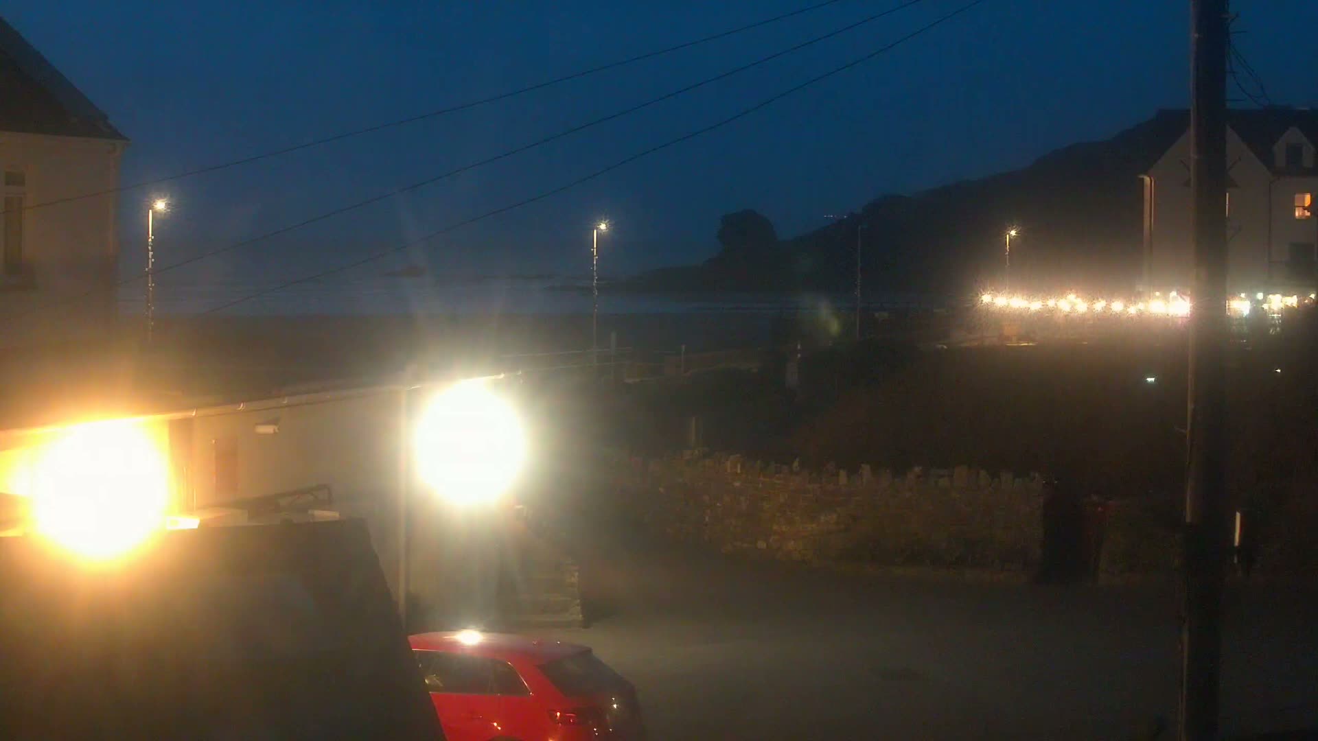 At night, bright lights illuminate a seaside town scene with a red car parked near buildings and a low stone wall, under a dark, clear sky.