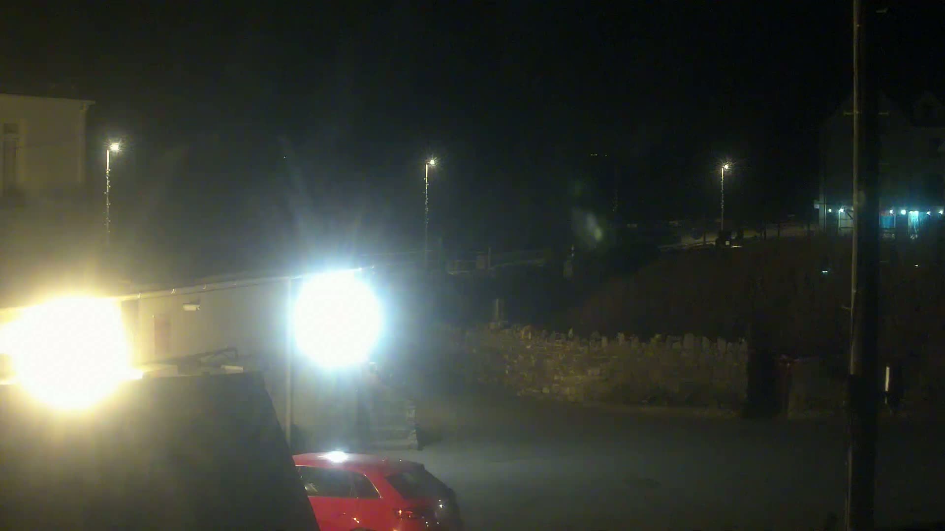 A nighttime outdoor scene shows a red car parked near a building, illuminated by bright headlights and streetlights.