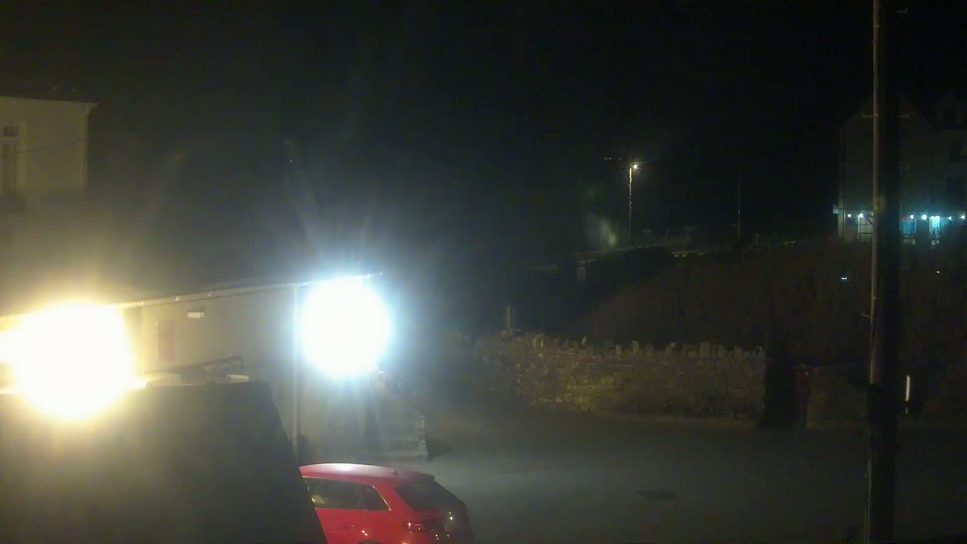 A nighttime, dimly lit scene shows a red car parked near a building and a low stone wall under bright artificial lights.