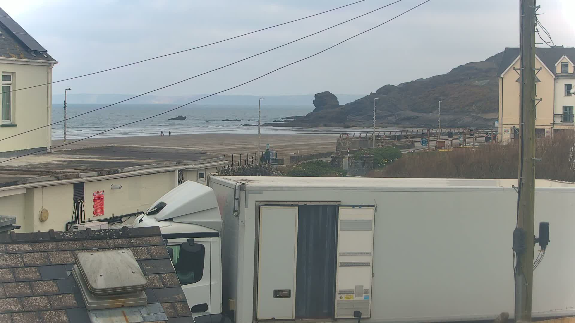 A view of a beach with a few people walking on it, seen from behind buildings and a large white truck, under an overcast sky.
